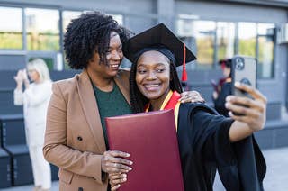 A female graduate wearing a cap and gown and holding a diploma takes a selfie with an older woman