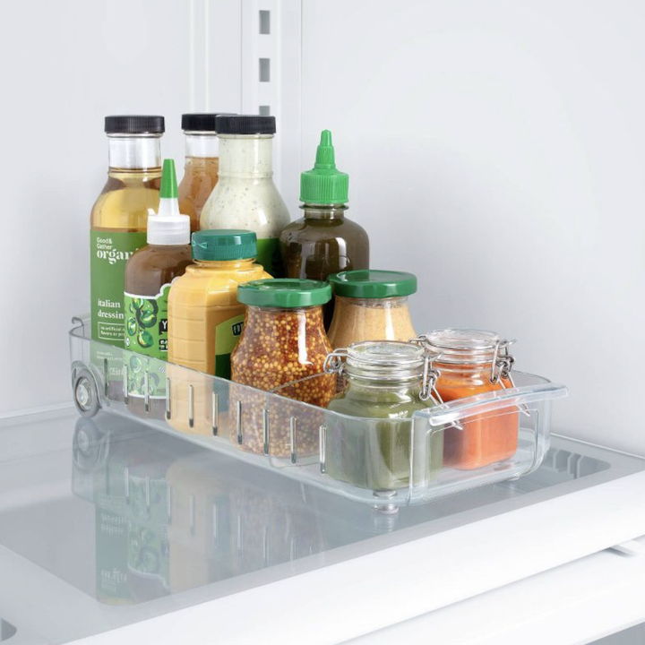 clear roll-out fridge caddy stocked with condiments on a white fridge shelf