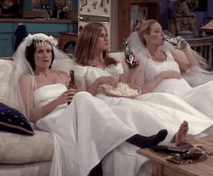Monica, Rachel, and Phoebe drinking and sitting on the couch in wedding dresses