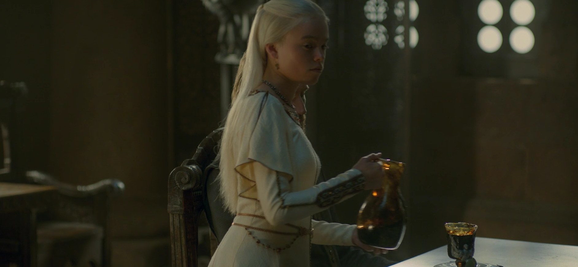 Rhaenyra stands besides a table posed to pour a drink into a cup