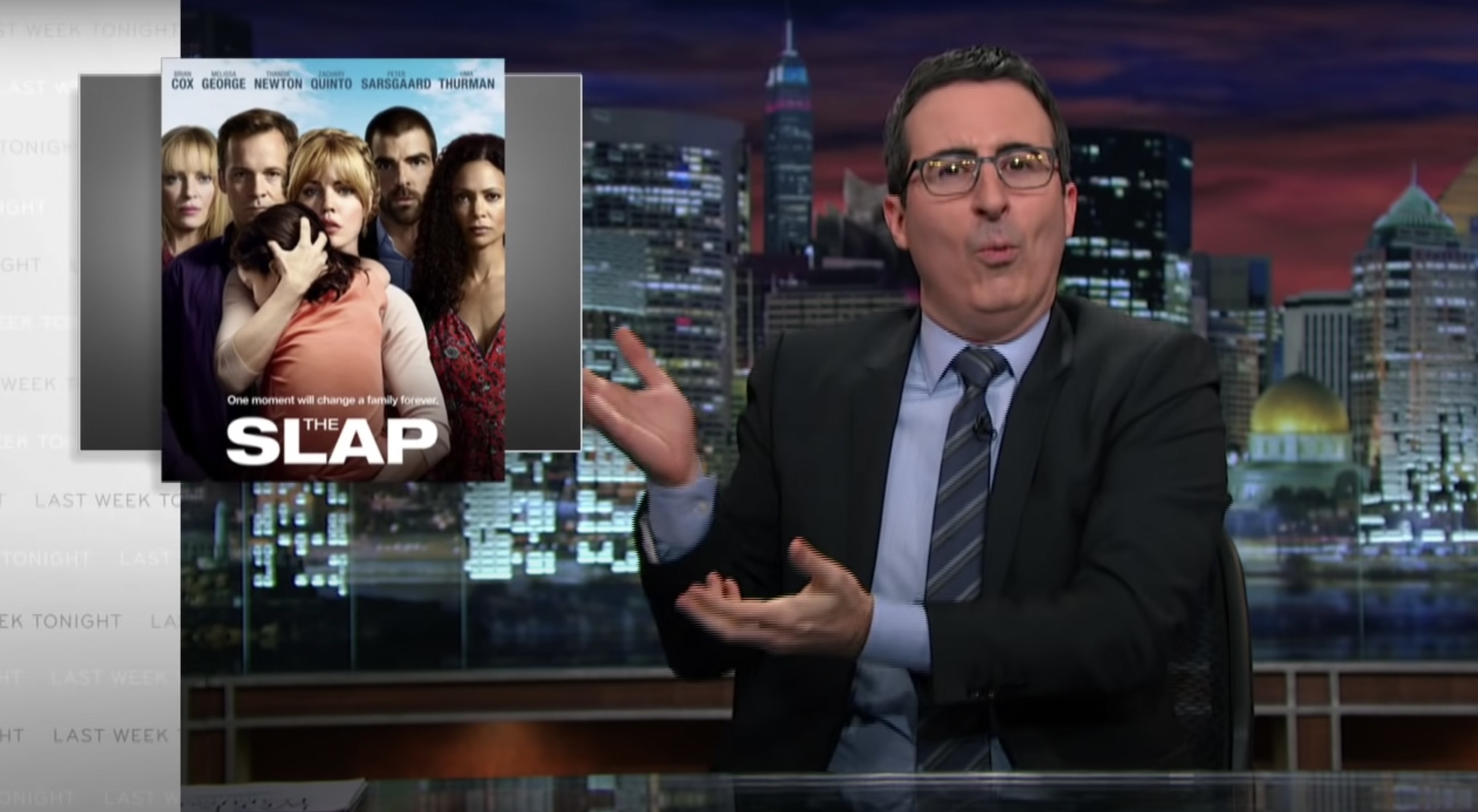 John Oliver at his desk, gesturing in confusion to an image of the tv show The Slap