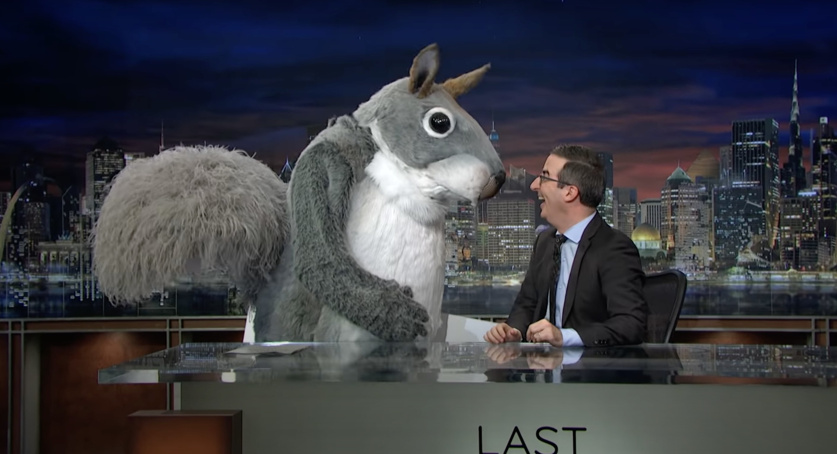 John Oliver at his desk smiling at a giant squirrel mascot standing next to him