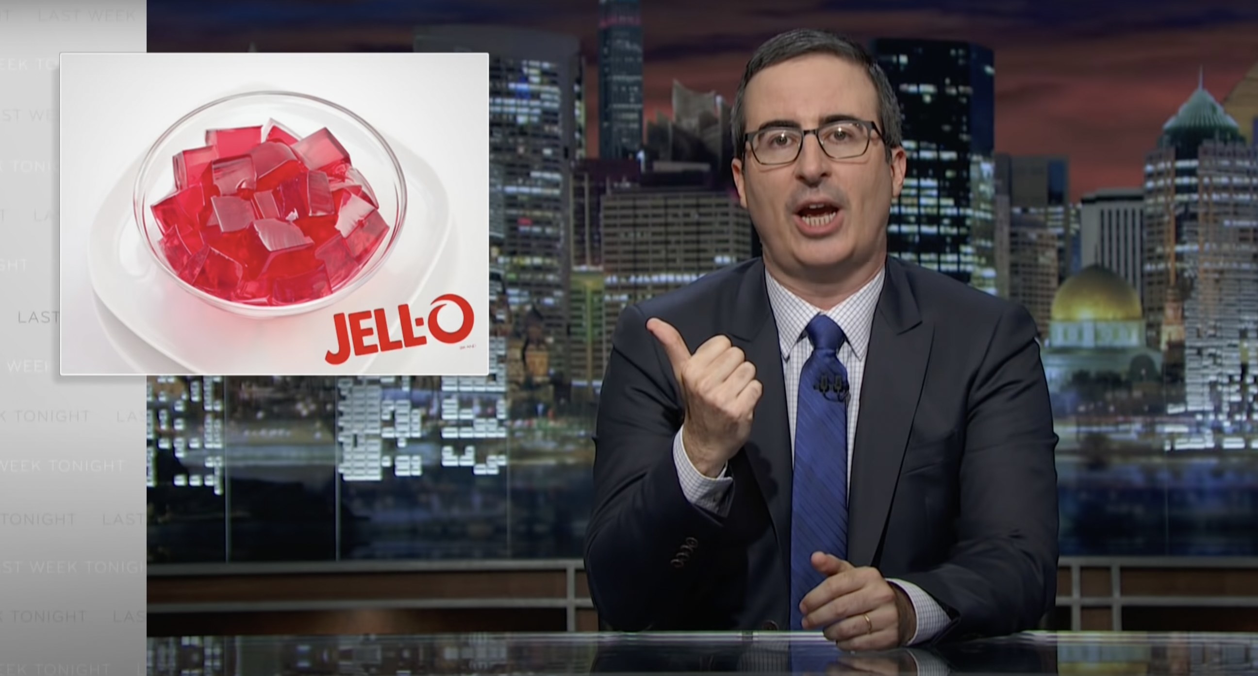 John Oliver at his desk, pointing to an image of Jello next to him