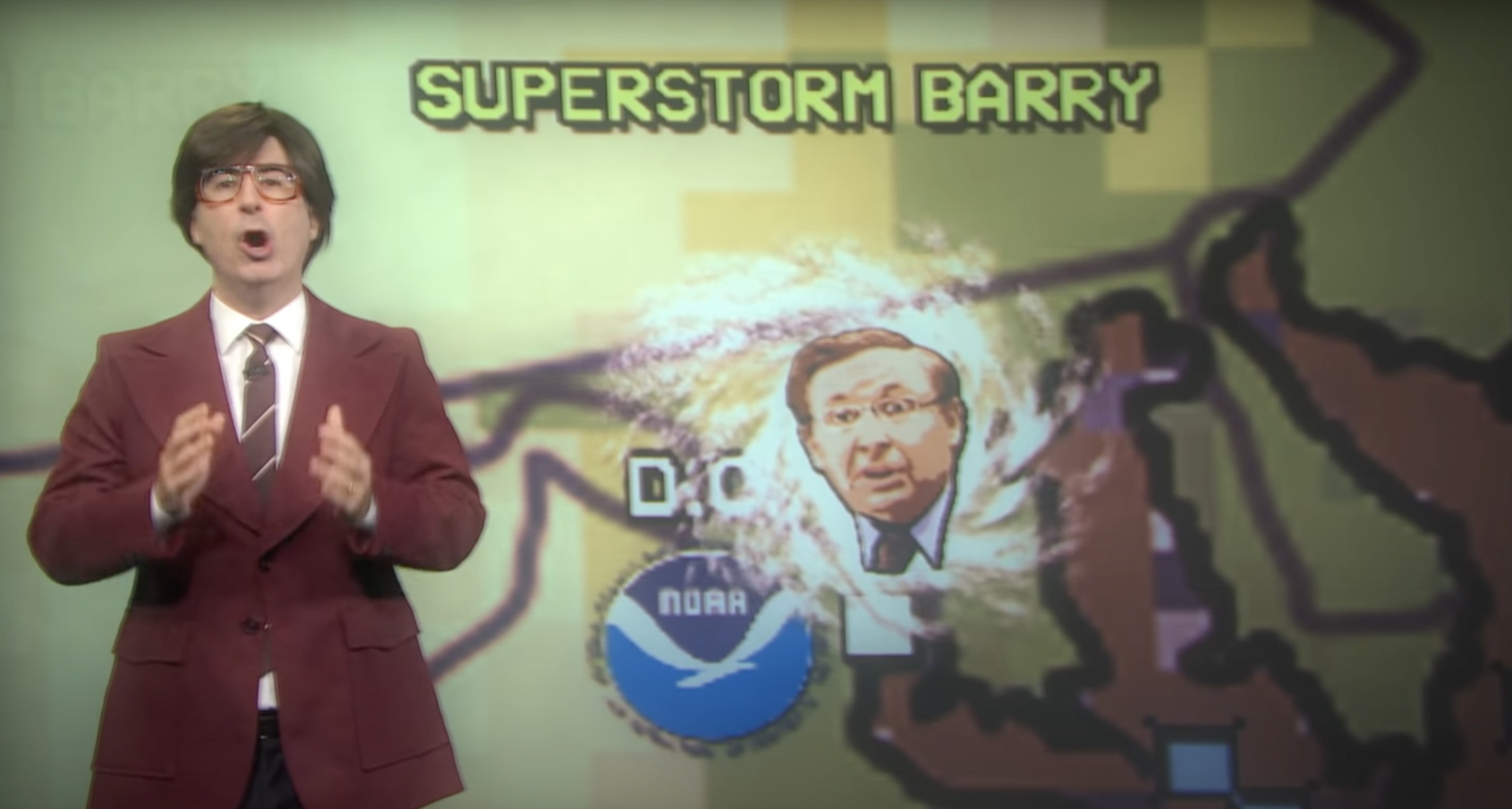 John Oliver wearing a wig, dressed as a 70s meteorologist, in front of an image of a superstorm Barry over Maryland, which is a hurricane with the face of someone named Barry on it