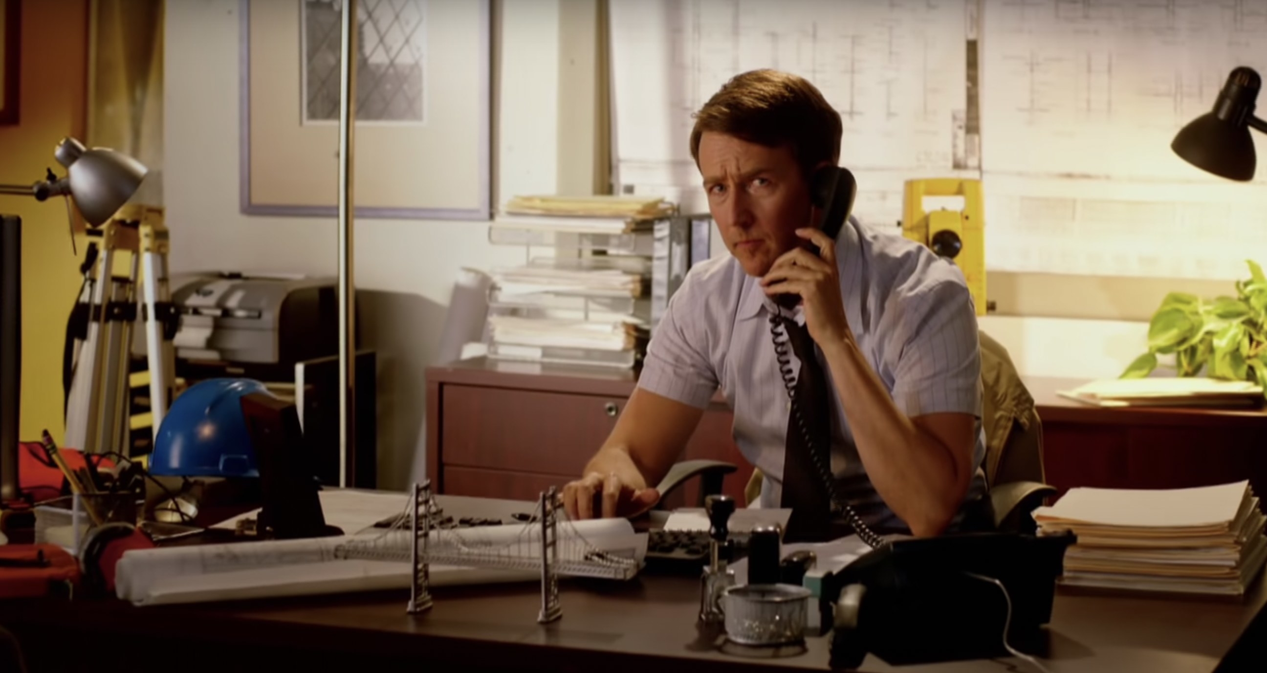 Edward Norton sitting at a desk on the phone, looking at the camera