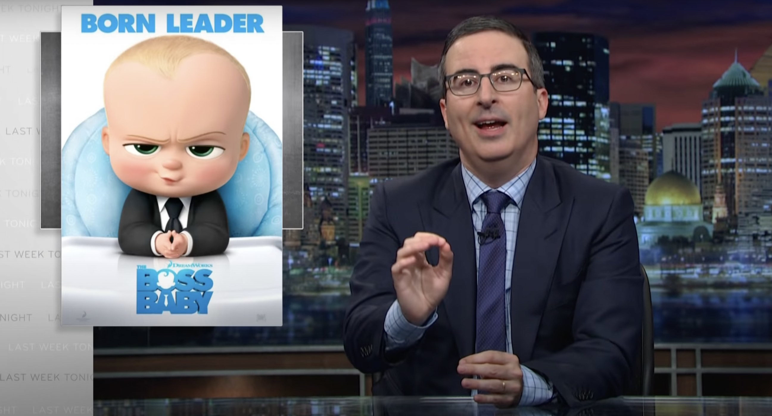 John Oliver at his desk with an image of the Bossy Baby poster next to him