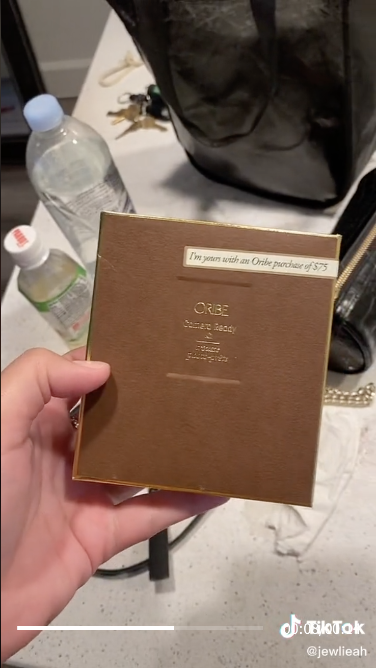 Julia holds an &quot;Oribe cCamera Ready&quot; card with &quot;I&#x27;m yours with an Oribe purchase of $75&quot; on it