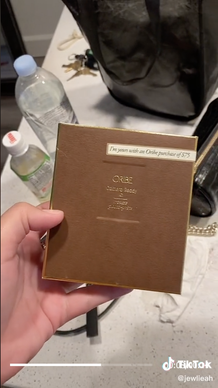 Julia holds an "Oribe cCamera Ready" card with "I'm yours with an Oribe purchase of $75" on it