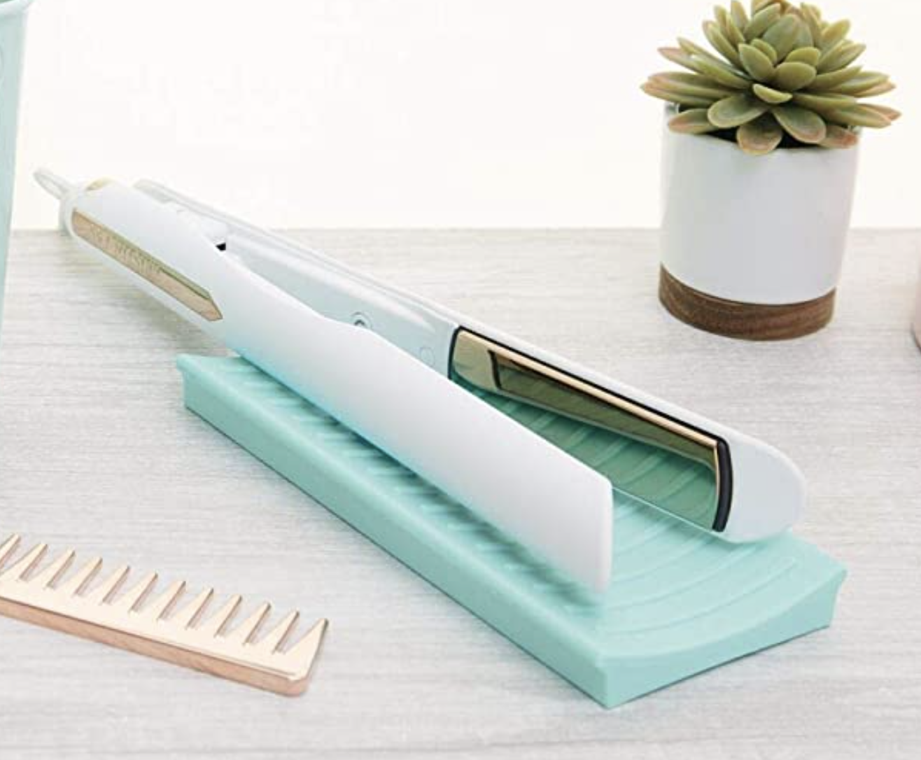 The heat mat with a flat iron on it, next to a plant and a metal comb