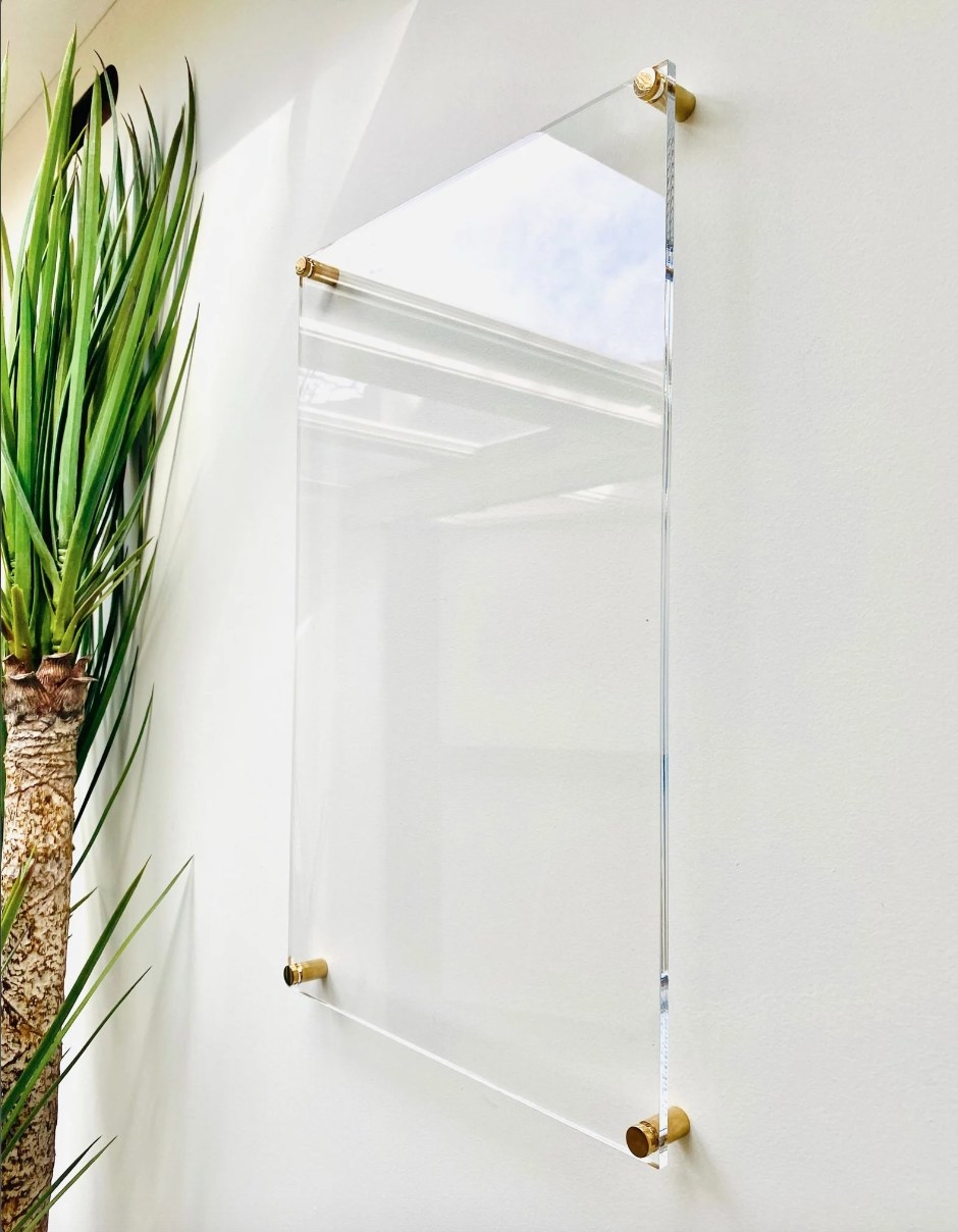 the clear sheet of acrylic floating on gold pegs just above a white wall with a plant next to it
