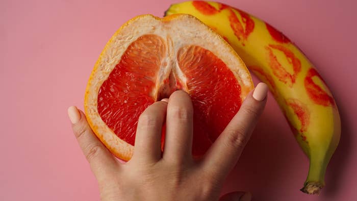 A hand with two fingers inserted into the center of a halved grapefruit, next to a banana with lipstick kisses on the peel