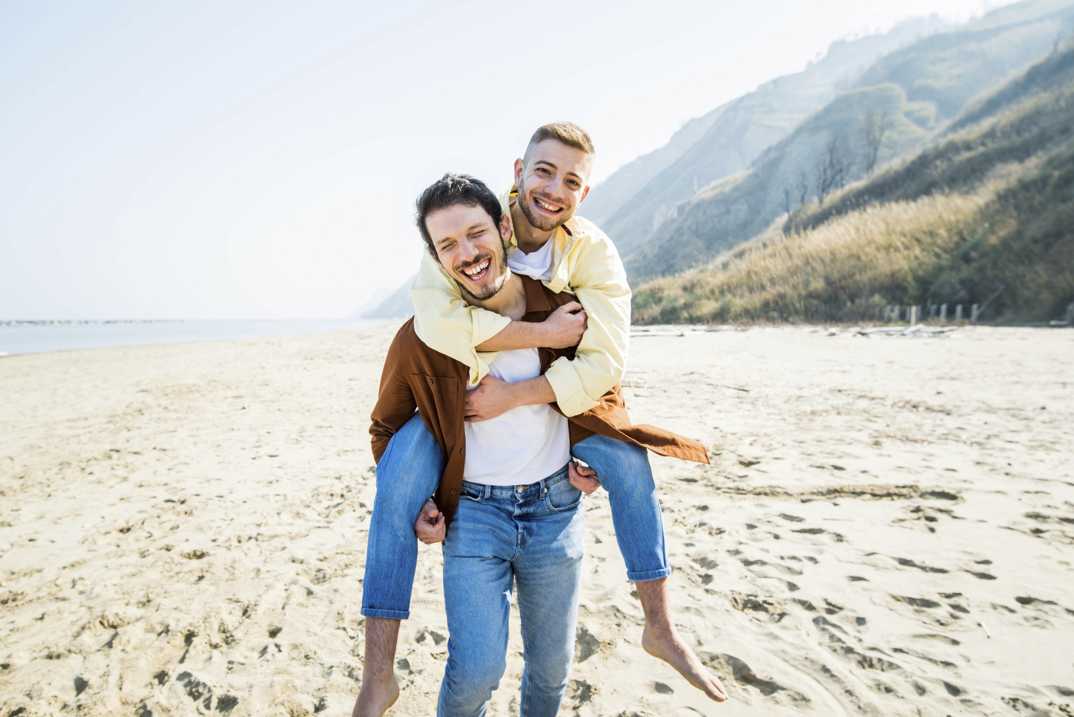Gay couple laugh on the beach as one man gives the other a piggy-back
