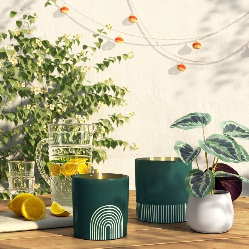 the green candle with an arch design burning on a table next to pitcher of lemon water and plants