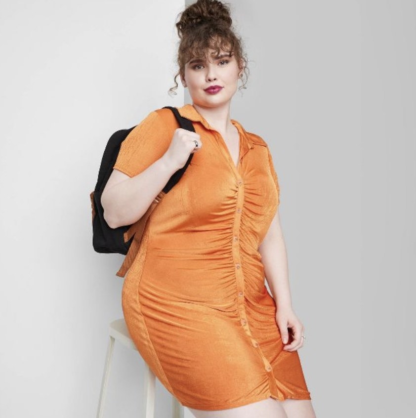 The model wears the orange ruched dress while sitting on a chair