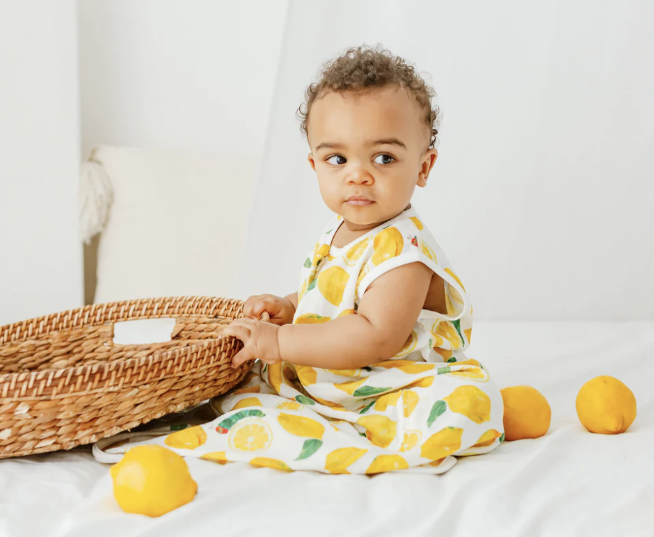 A baby sitting in a bed wearing a lemon print sleep sack