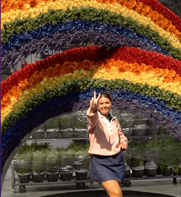 Jesse stands below flower rainbow sculpture. Gives peace sign and kicks up heel.