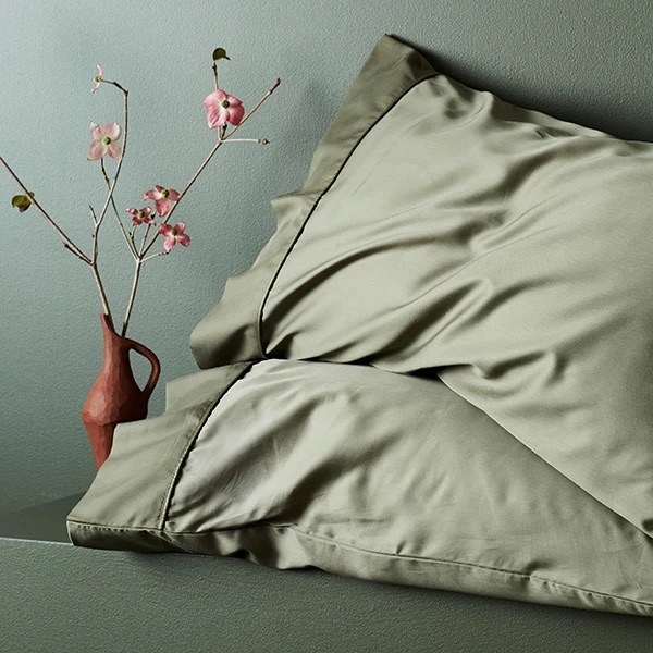 A pair of jade colored pillowcases in a soft silk sitting next to sprigs of cherry blossom in a funky vase