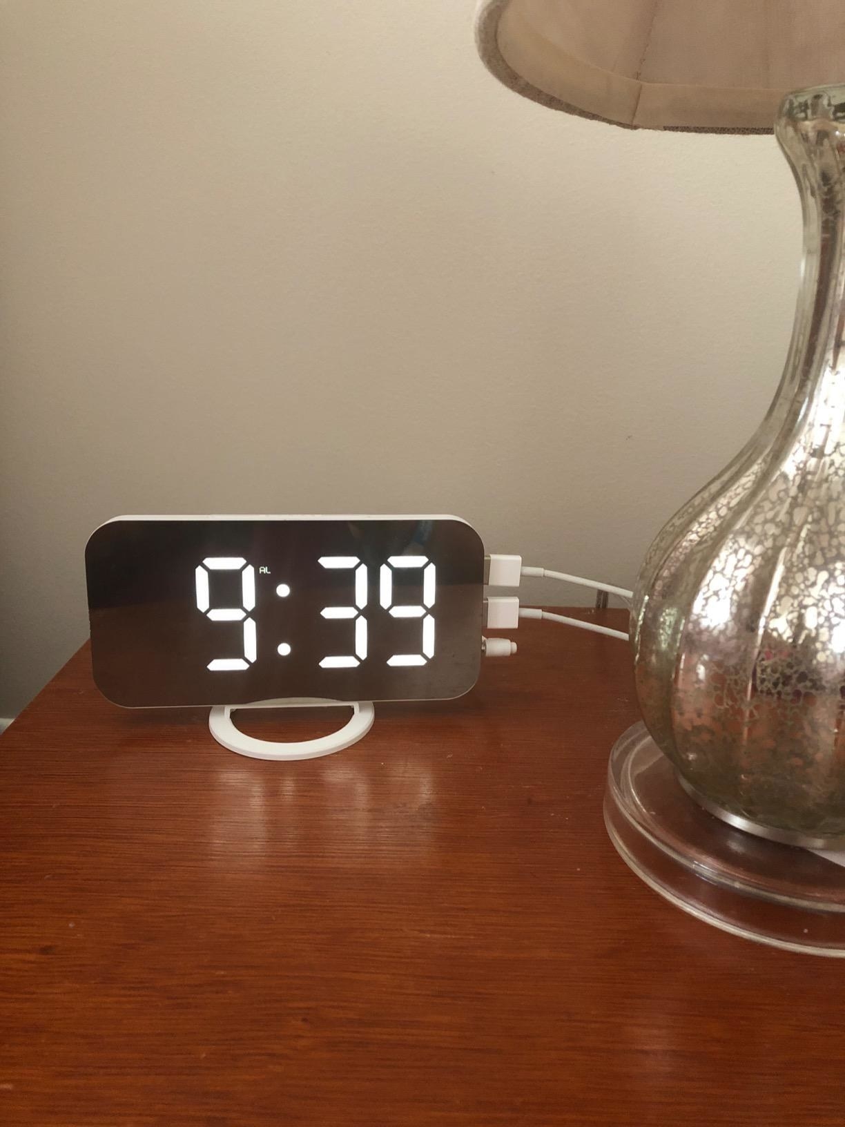 a review photo of the silver mirrored clock next to a lamp on a nightstand