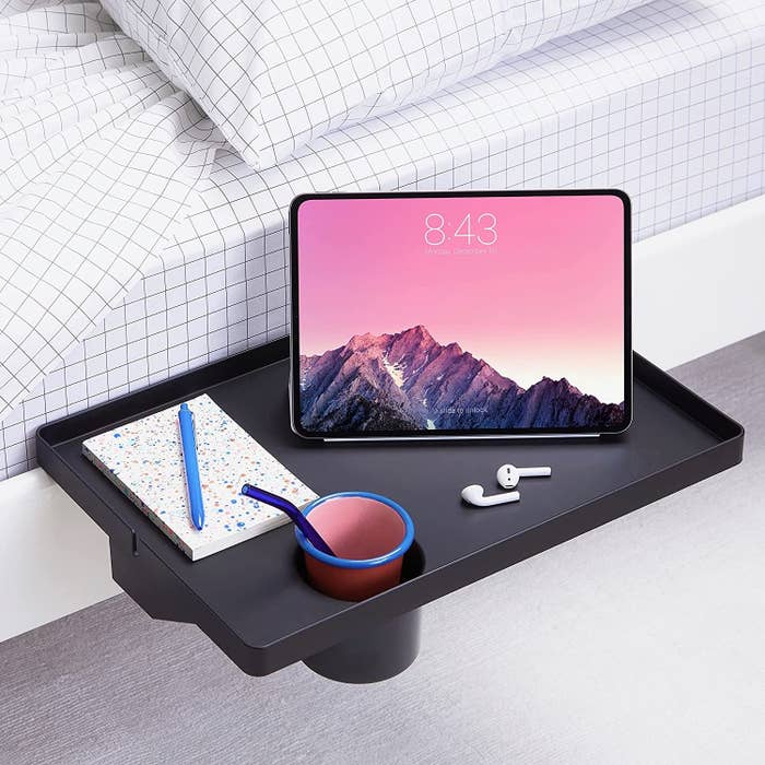 Bedside tray with a notebook, pen, bowl, wireless earbuds, and a tablet displaying a upland scene. Modern and organized setup