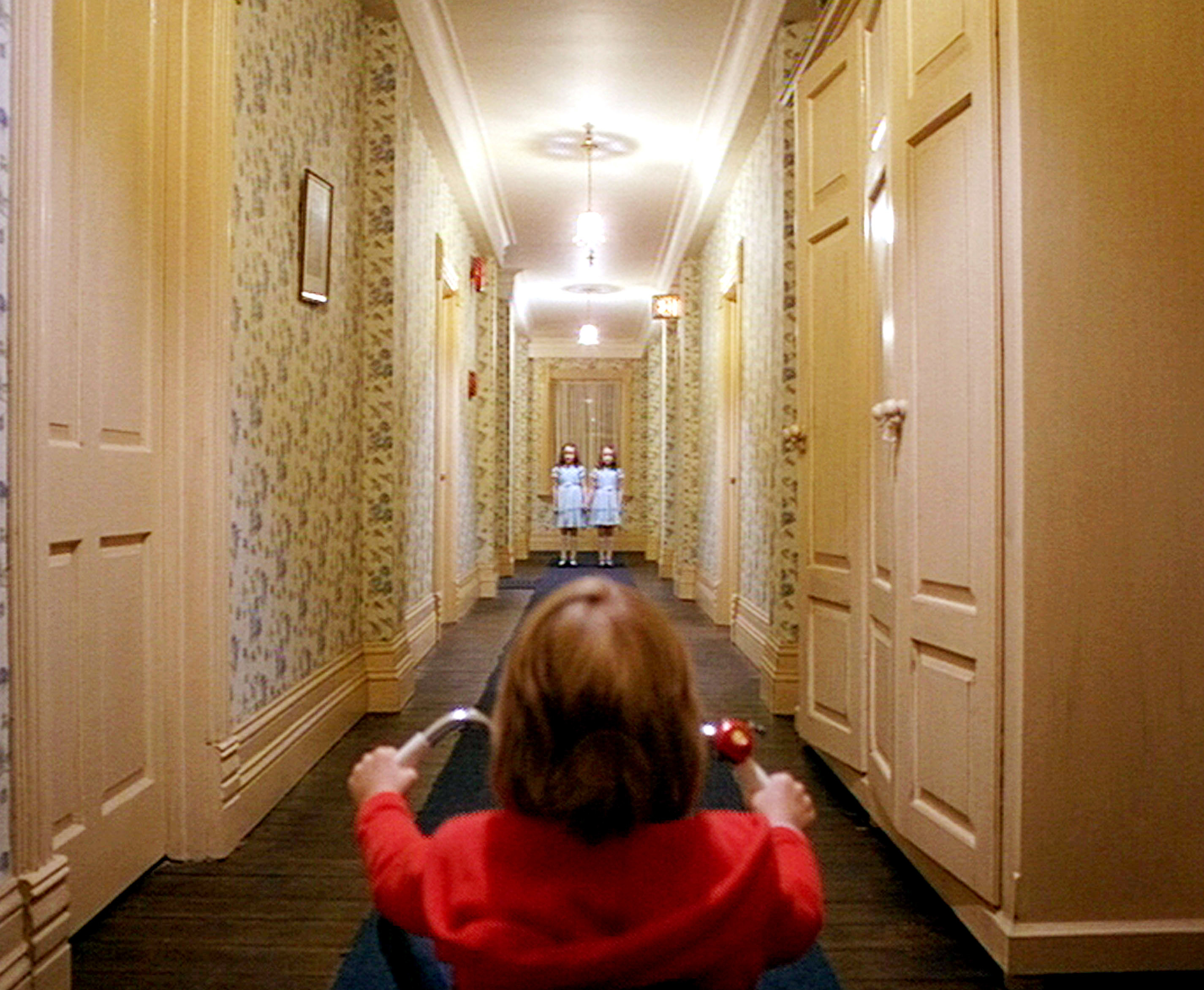 Twin girls standing at the end of a hallway and a little boy on a kids bike facing them