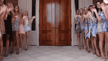 Young women waving outside of a sorority house as its doors open