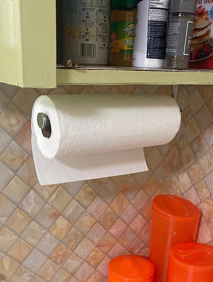 Reviewer&#x27;s silver paper towel holder mounted under kitchen cabinet with roll of paper towels on it