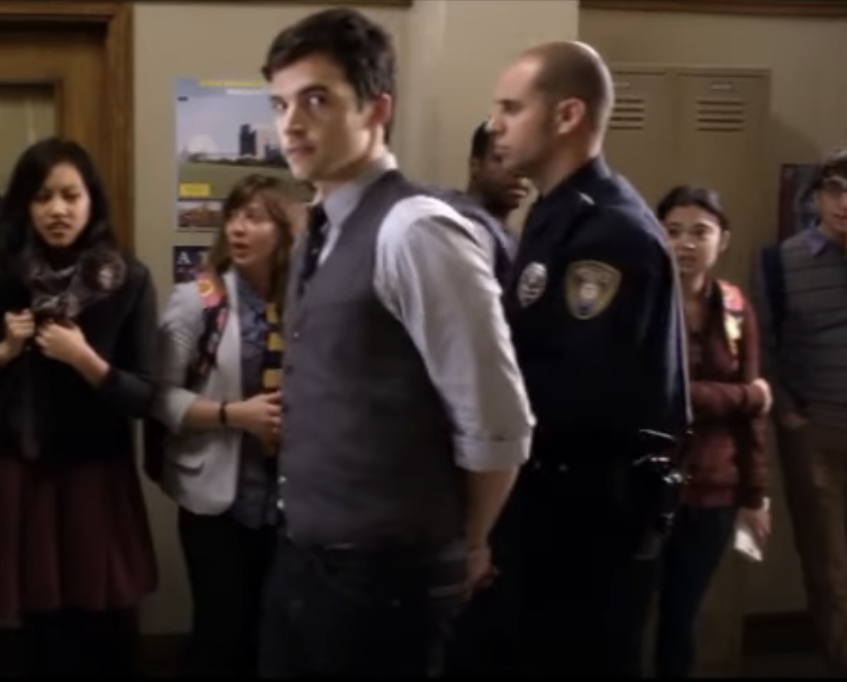 Ezra in a school hallway with a cop as people look on