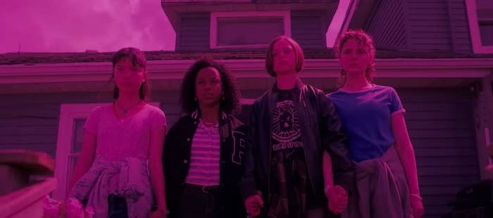 The four paper girls stand in a row holding hands and staring ahead in front of a house; everything is bathed in a pinkish purple light