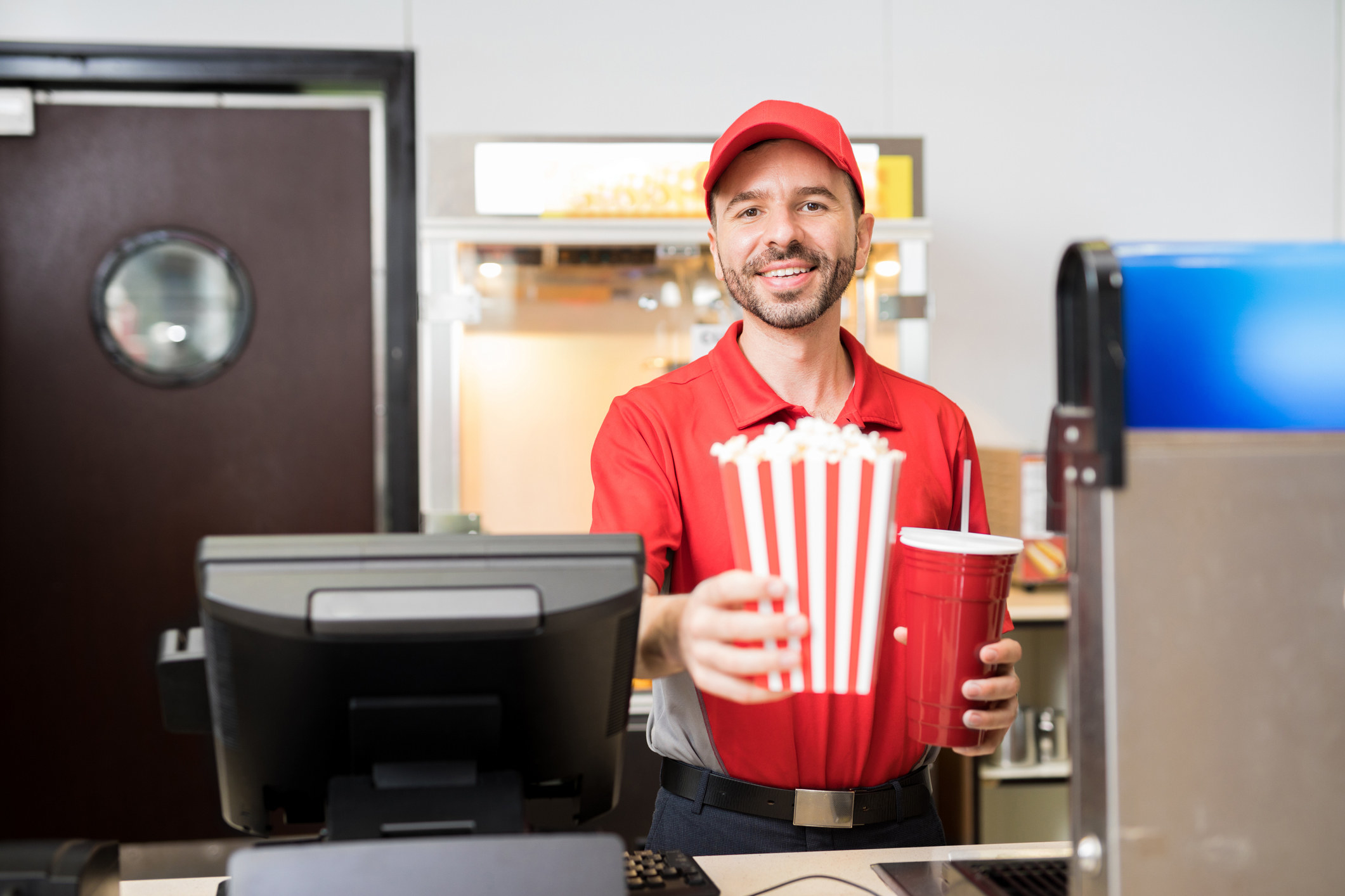 Movie theater worker behind the counter holding a large beverage cup with straw and popcorn