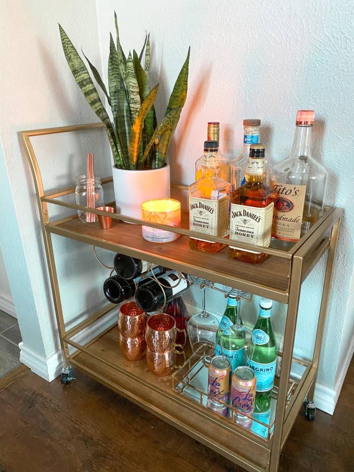 Reviewer&#x27;s photo of the gold cart storing different types of alcohol, wine bottles, mule mugs, wine glasses with additional decorative objects on top shelf