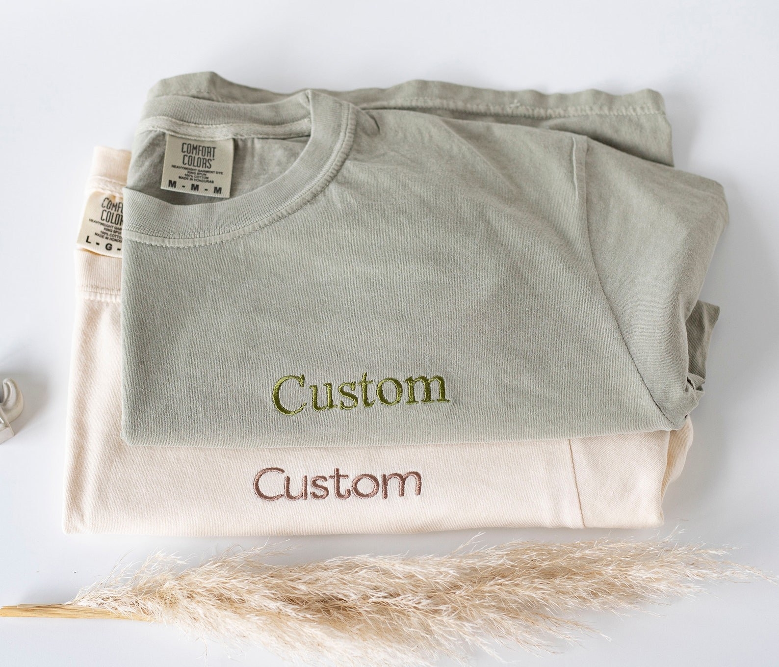 two t-shirts in beige and light green with the word &quot;custom&quot; embroidered on them