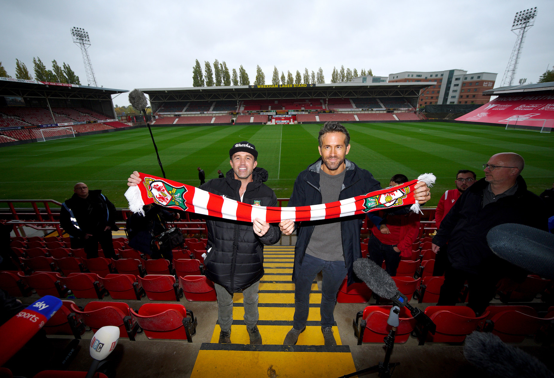 Rob and Ryan holding up a striped team scarf at a soccer stadium