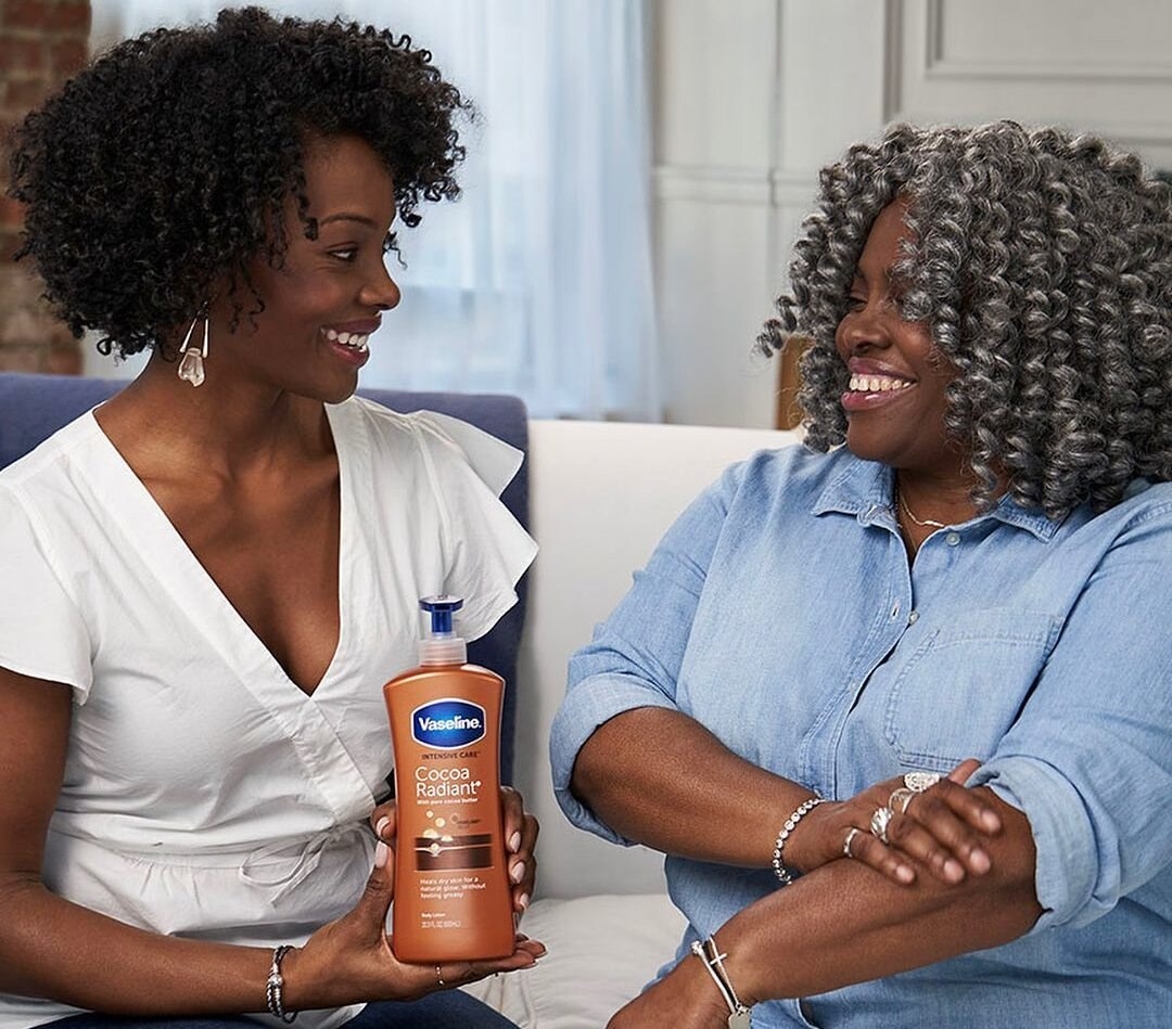 Two people smiling while one is holding the body lotion and the other is rubbing it on their arm