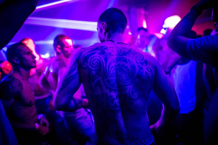 Shirtless men dance under purple lights, with one man&#x27;s back and large tattoo directly in front of the camera