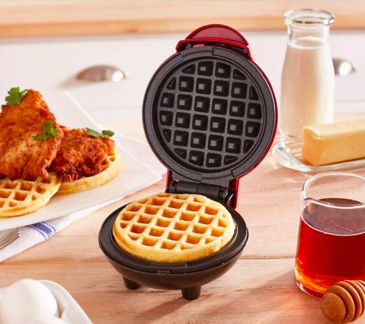 Red waffle maker with a cooked waffle inside next to a plate of fried chicken and waffles and a glass with honey