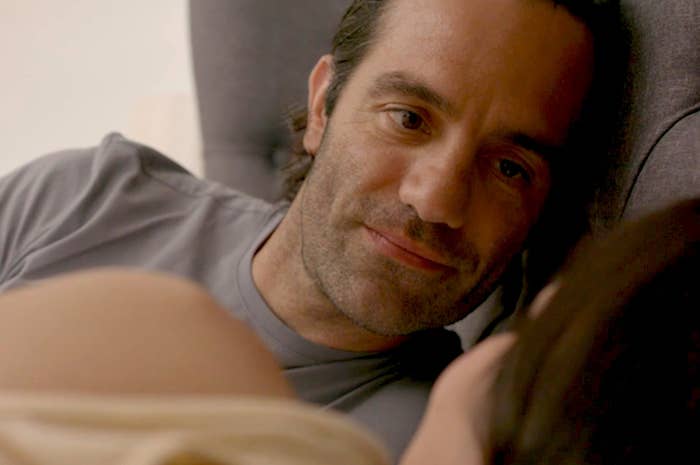 A man lies in bed looking at a woman who has her back to the camera and is out of focus
