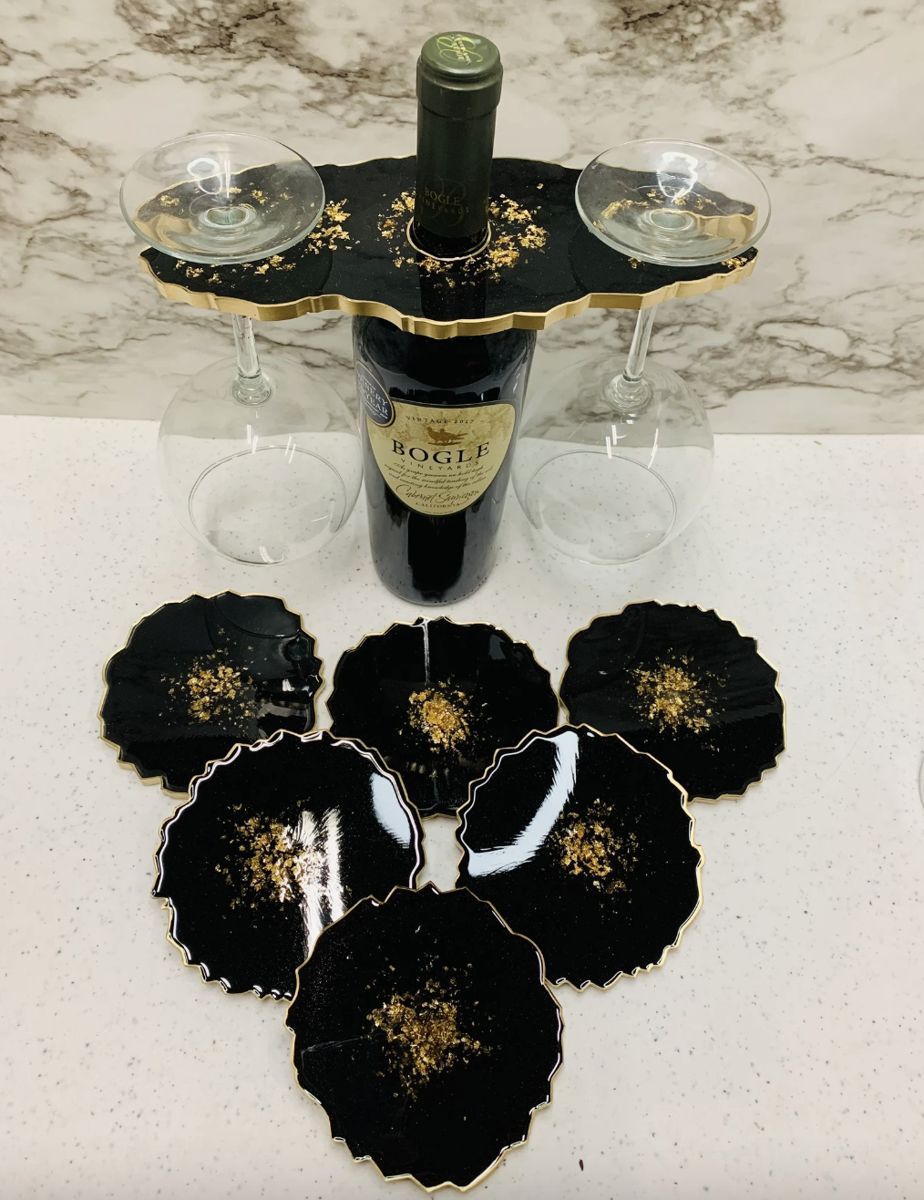 black geode coasters with gold flecks in the center and a gold rim next to a matching wine caddy that&#x27;s holding a bottle of wine and two wine glasses