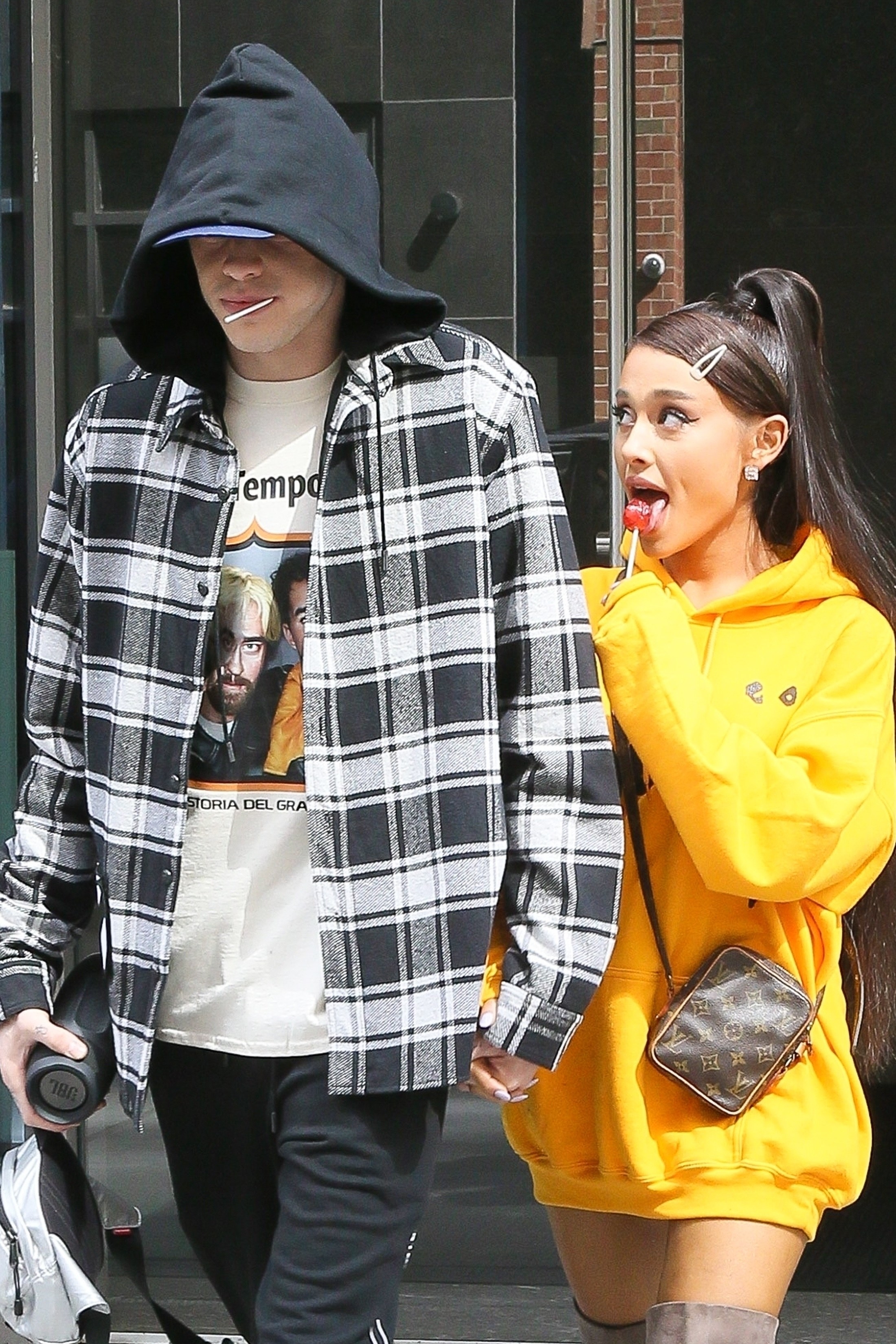 Pete and Ariana walking hand in hand; Pete has a lollipop in his mouth, while Ariana is licking one of her own