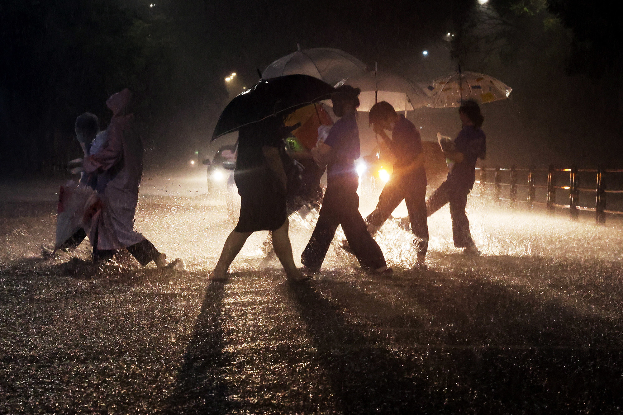 People wearing rain coats and holding umbrellas are silhouetted by car headlights as they cross a street in heavy rain