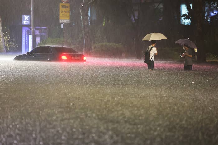 Two people stand in knee-deep water under umbrellas, looking at a nearby parked car whose brake lights are on