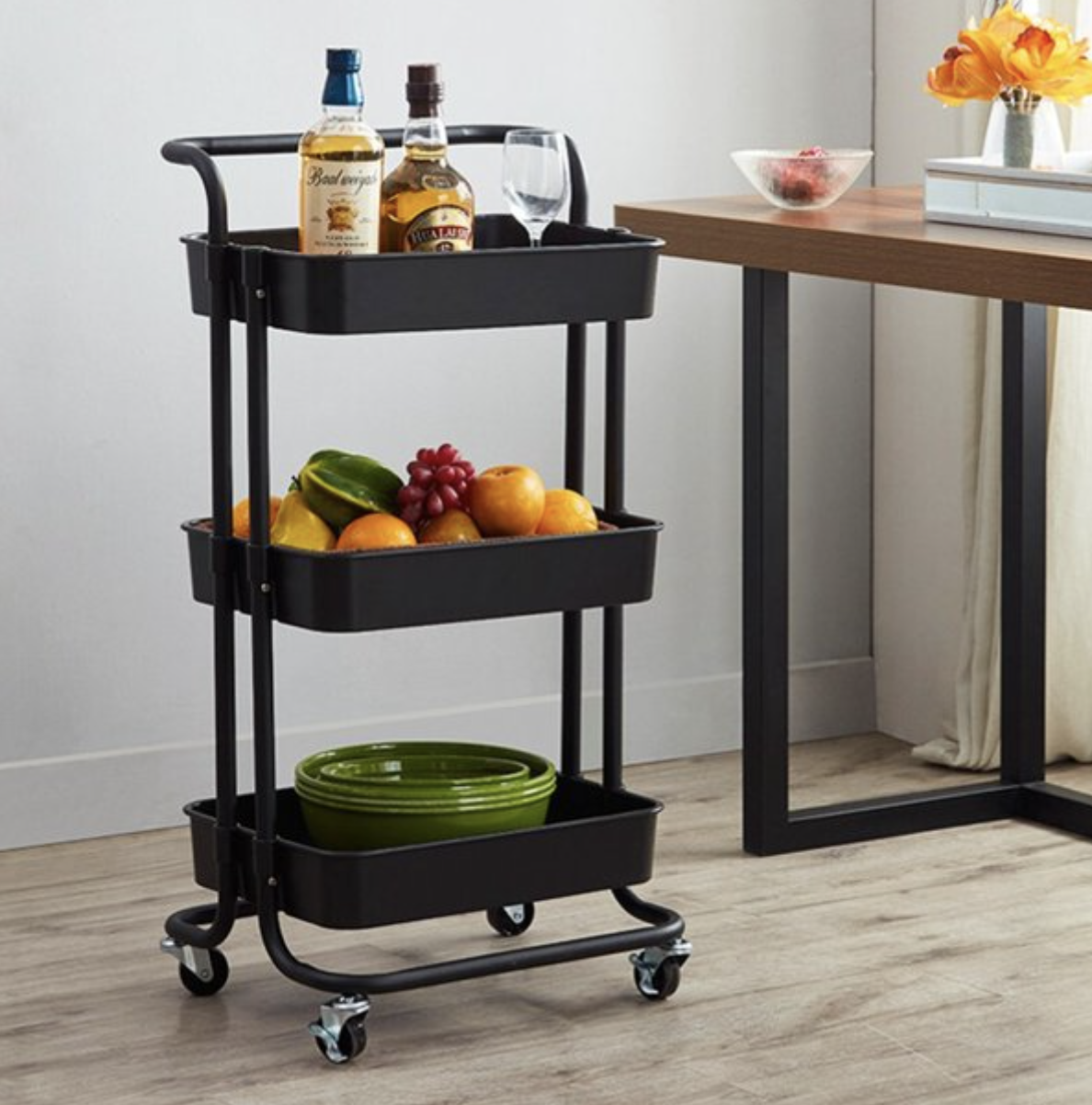 black three-tier rolling cart with shelves filled with alcohol bottles, fruit, and green plates
