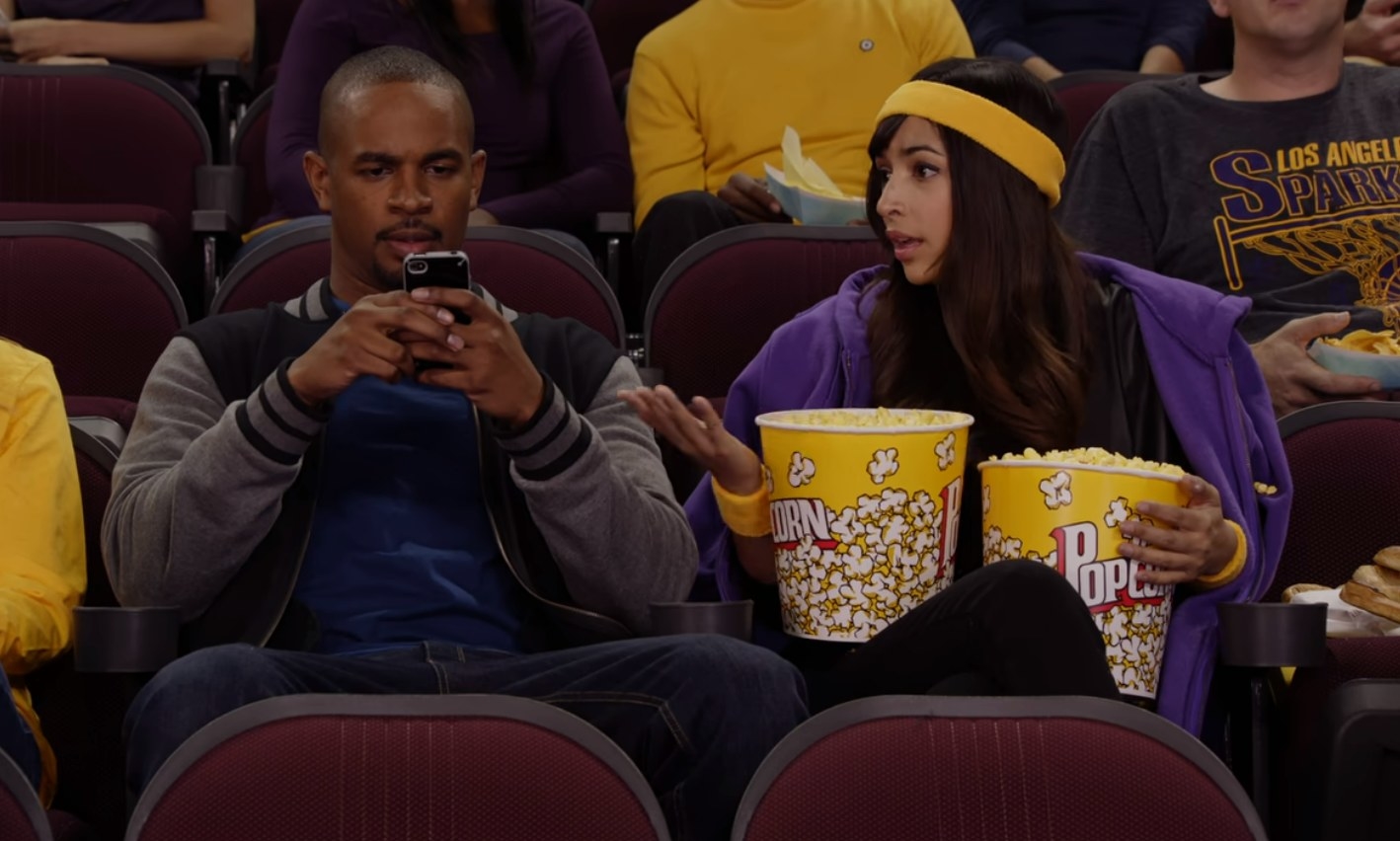Coach texting sitting next to Cece who is holding two buckets of popcorn.