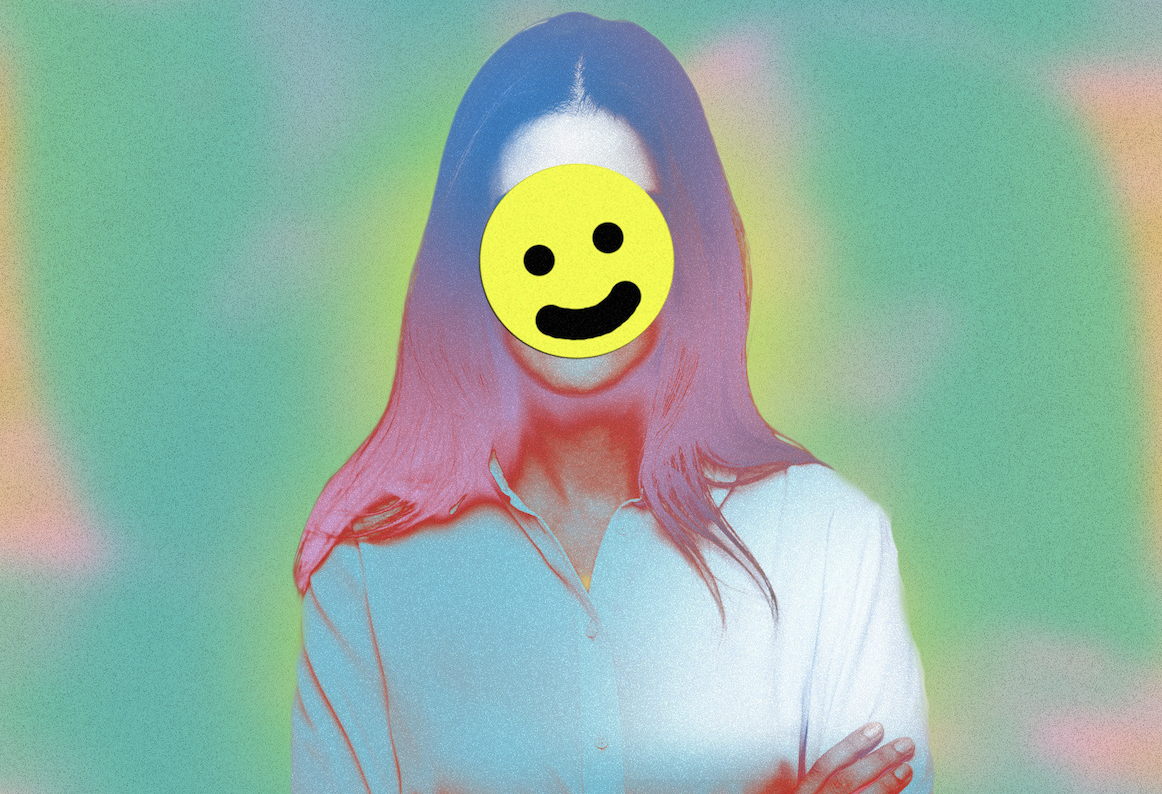 A person in a button-down and long hair faces the camera; their face is covered by a cartoon smiley face