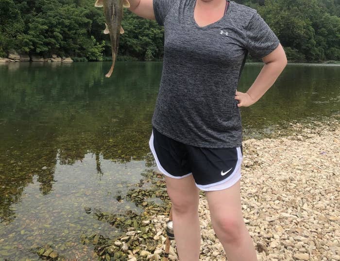 Reviewer standing by lake holding fish while wearing the grey heather v-neck with shorts