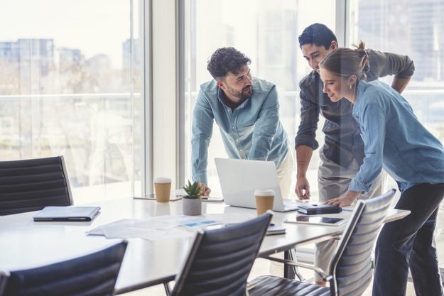 Two men and a woman look at a laptop together in their office&#x27;s conference room