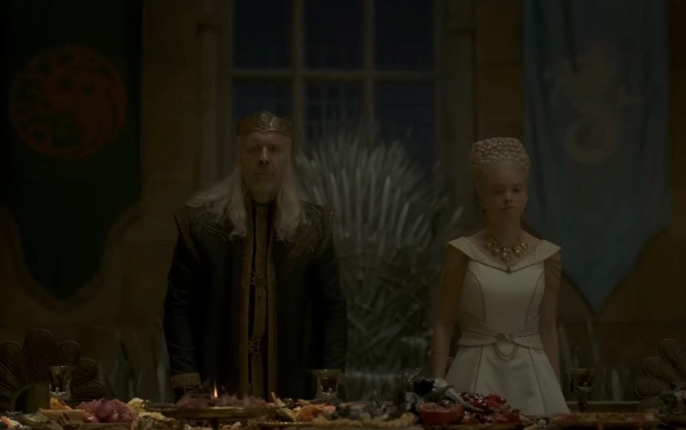 Viserys and Rhaenyra are dressed in formal attire in the throne room with a feast in front of them and the Targaryen and Velaryon banners behind them