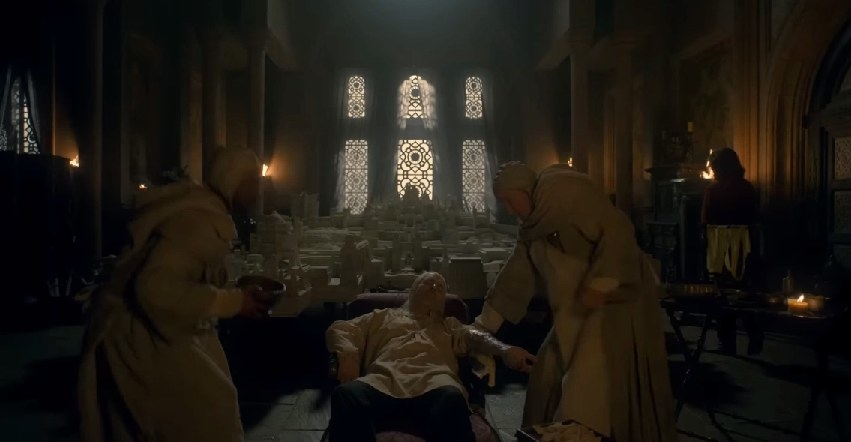Viserys is attended to by two maesters as he reclines in his chambers with an infected arm