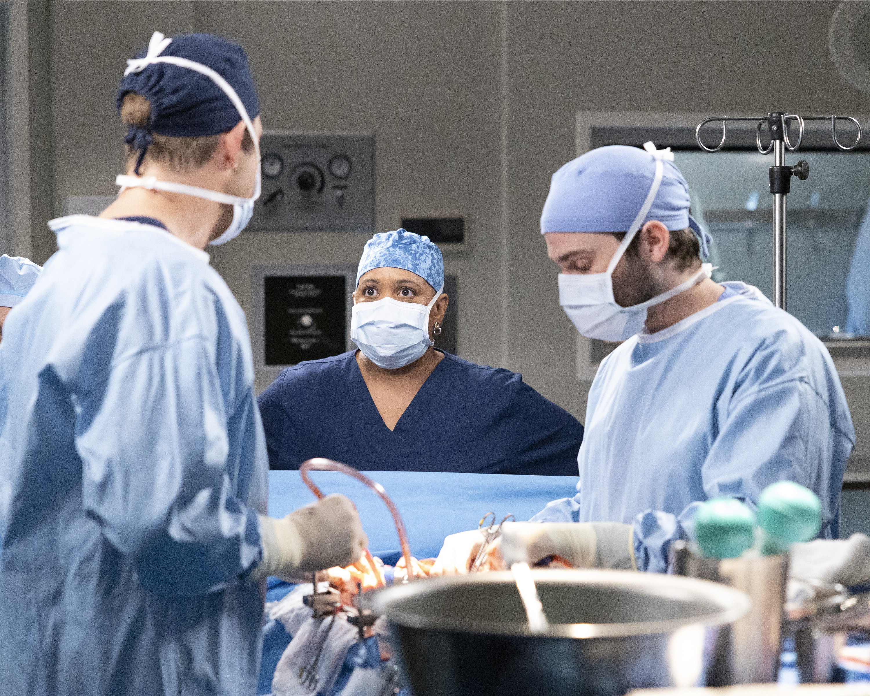 Bailey stands in the operating theatre wearing a mask and a scrub hat, while two men operate on a body