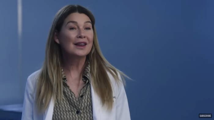 Meredith Grey, in a business shirt and lab coat, stands in a room talking to people