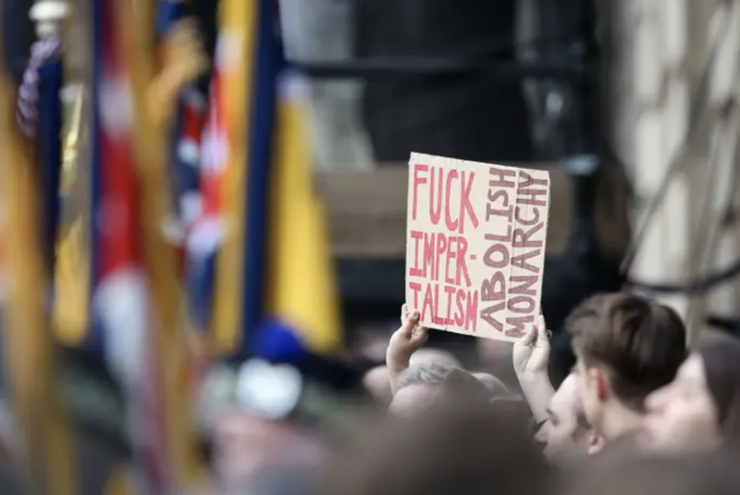 in a crowd, someone holds up a cardboard sign that reads &quot;fuck imperialism, abolish monarchy&quot;